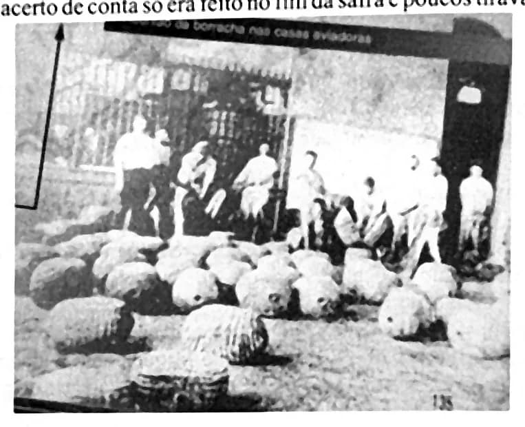 Fotografia antiga de pelas de borracha empilhadas em frente ao barracão de um seringal